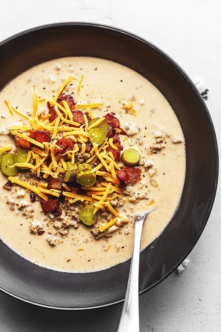 Bacon cheeseburger soup in a black bowl topped with sliced pickles, bacon and shredded cheese