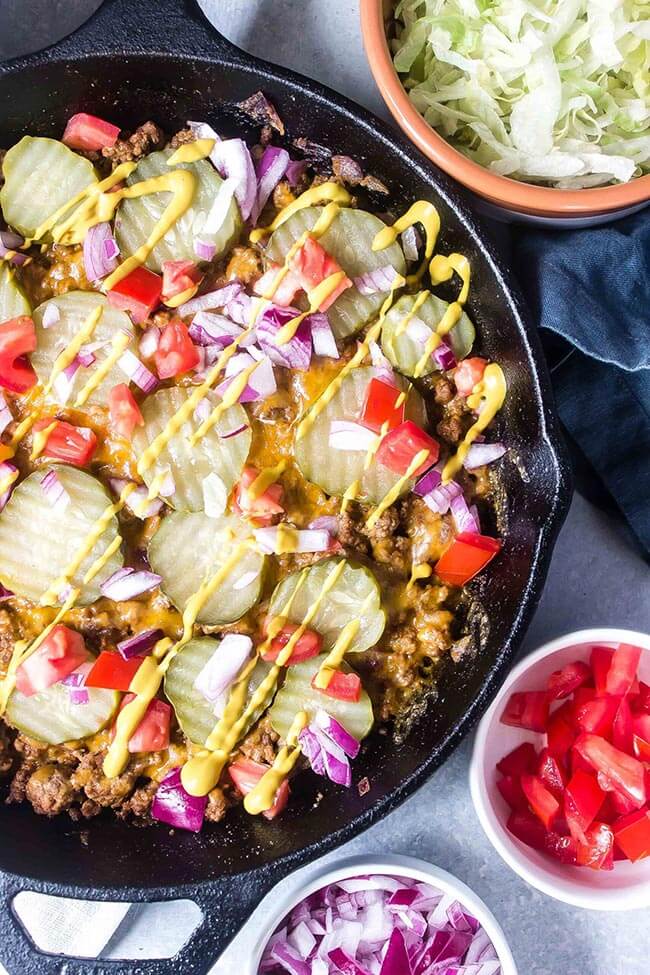 Cheeseburger cast iron skillet topped with melted cheese, sliced pickles and mustard drizzle.