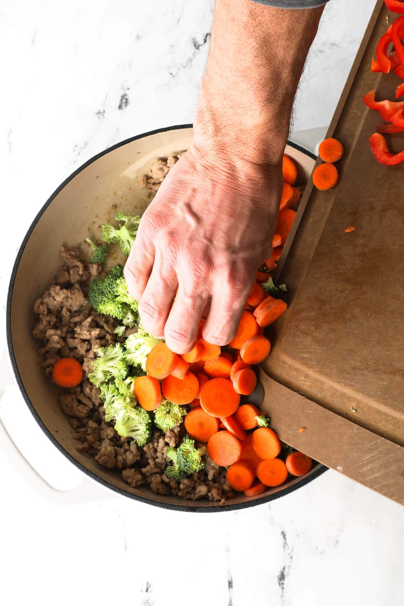 Adding sliced veggies to a skillet once the ground chicken is cooked.