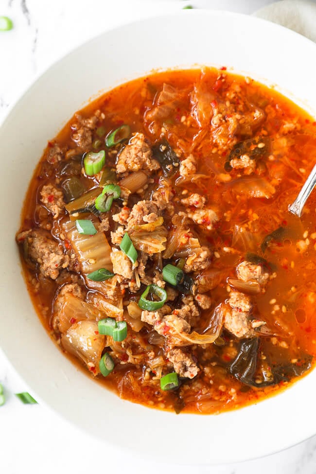 Overhead close up of kimchi soup in a bowl with a spoon in the bowl. Topped with chopped green onion. 