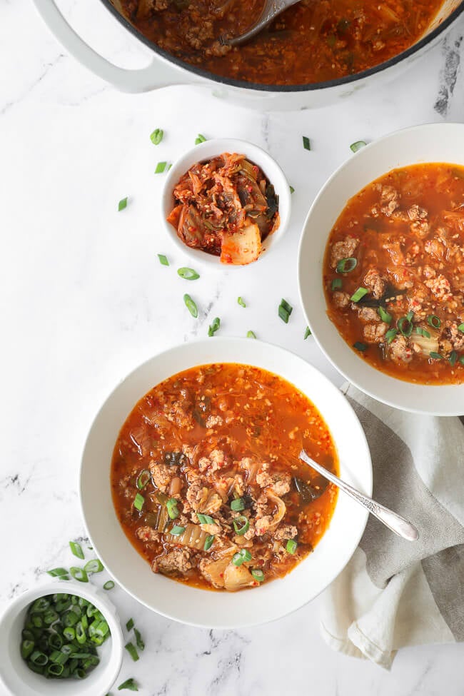 Overhead image of kimchi soup in soup pot and served up in two bowls with spoons in the bowls. 