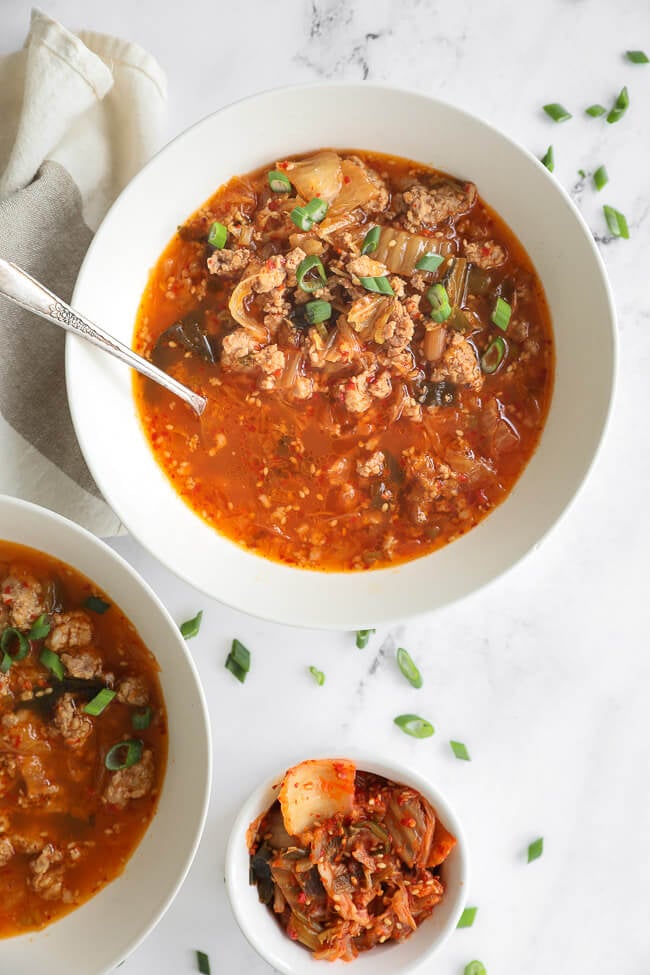 Overhead image of kimchi soup in a bowl with chopped green onion on top and a spoon dug into the bowl. 