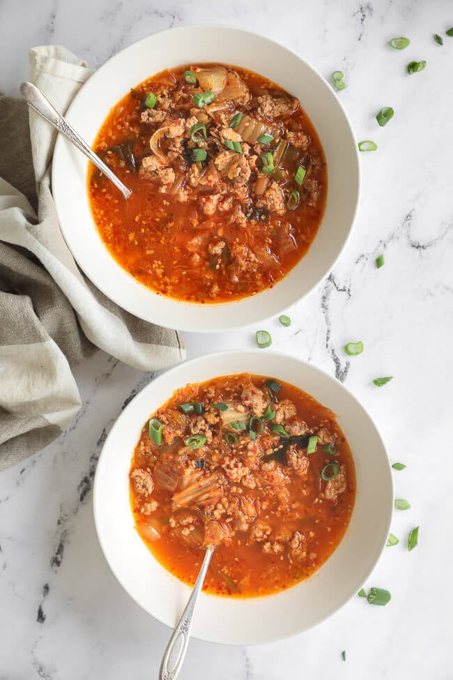 Overhead image of two bowls in a vertical line of kimchi soup with chopped green onion on to and spoons dug into the bowls. 