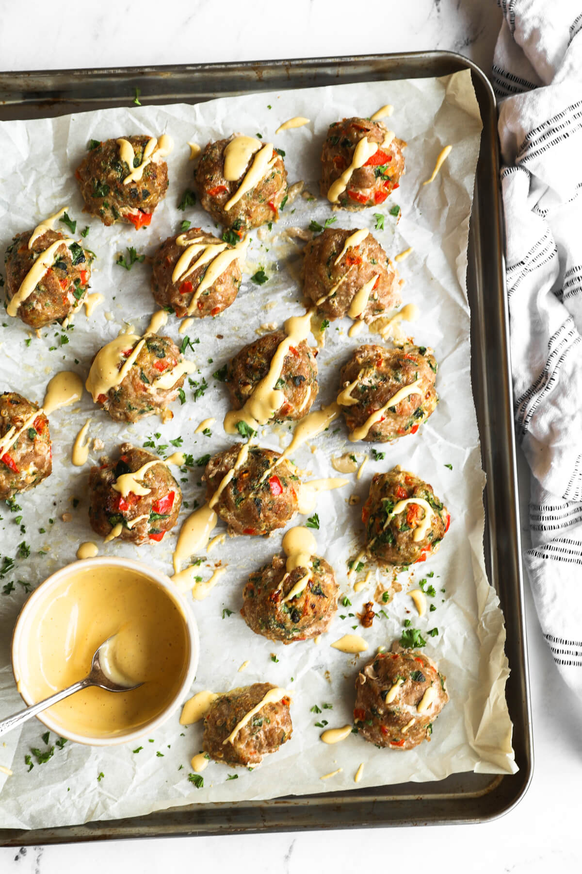 Overhead image of meatballs on a sheet pan with a side of spicy aioli and sauce drizzled on top of meatballs.