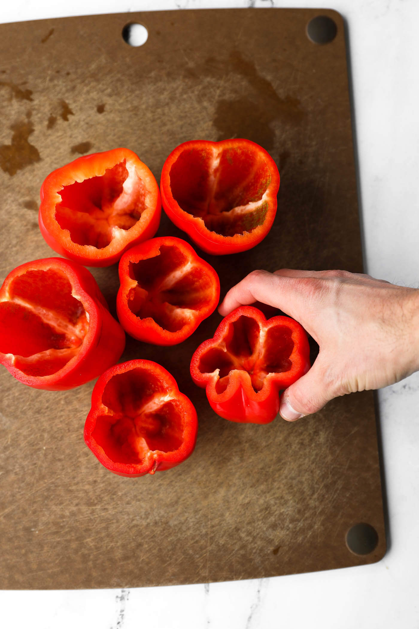 A hand placing a bell pepper on a cutting board. The bell pepper has the top sliced off and the seeds removed. There are 6 bell peppers on the cutting board.
