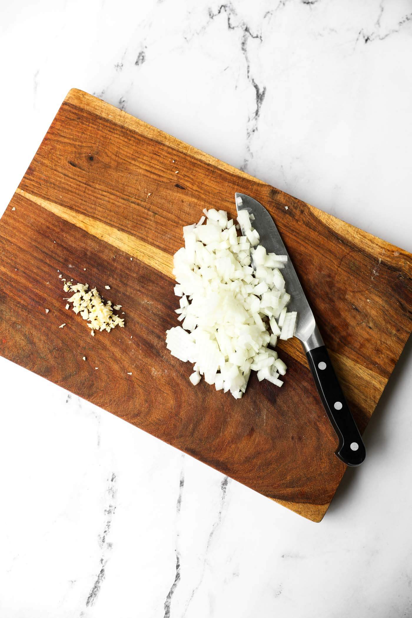 A cutting board with minced garlic and chopped onion on it with a knife partially covered by the chopped onion.
