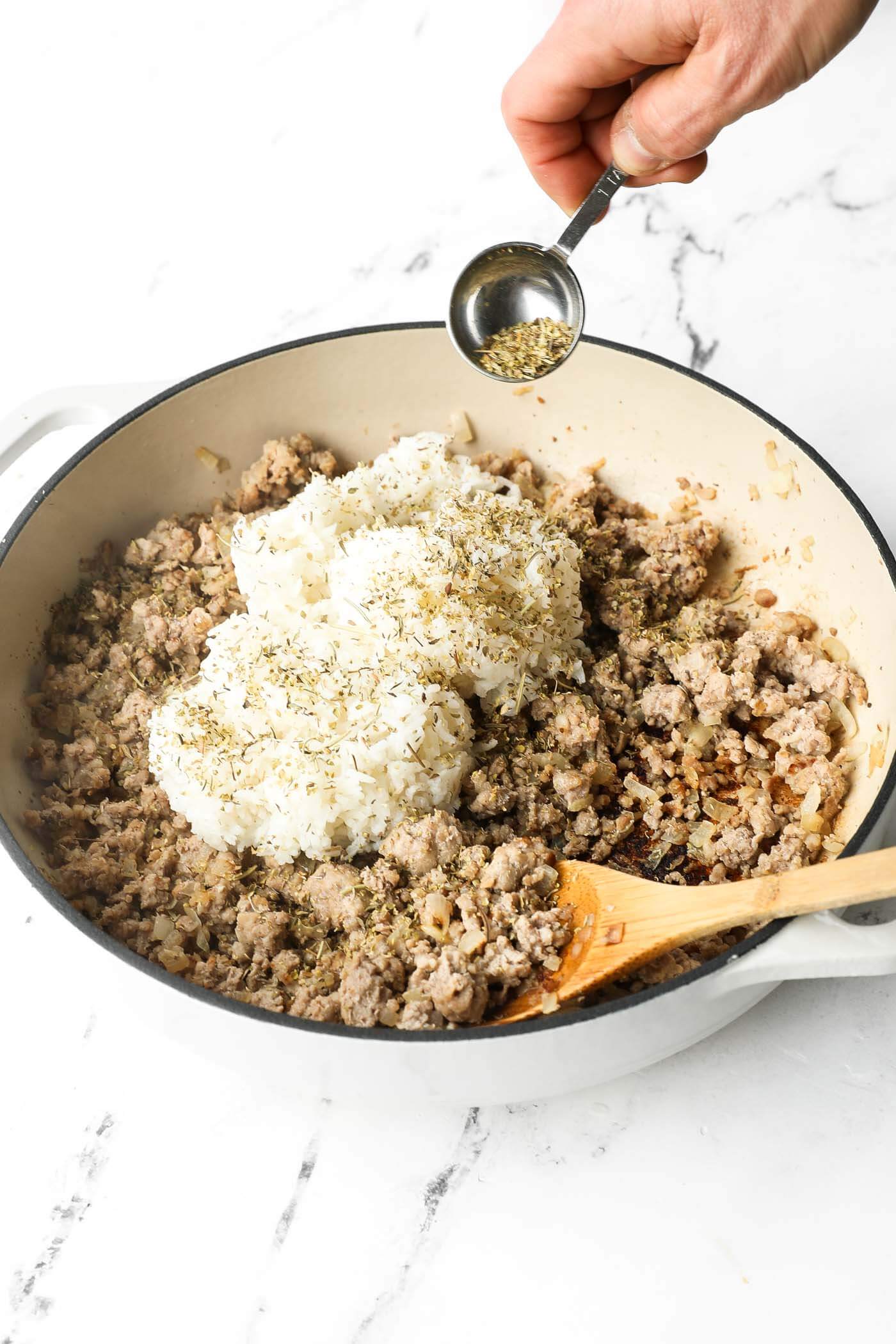 Angled image of a hand sprinkling Italian seasoning over the cooked ground pork and white rice in a skillet.