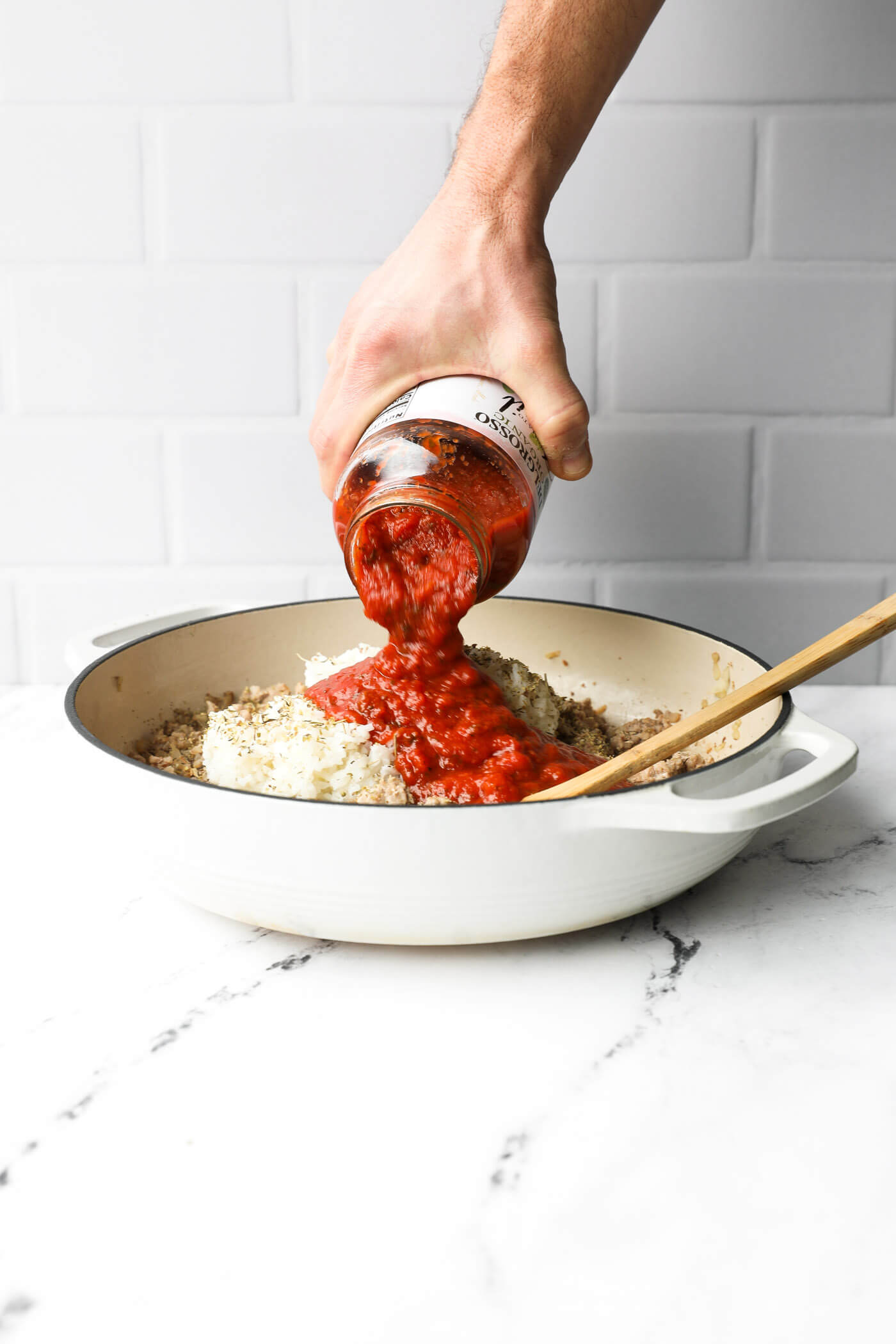 A hand pouring marinara sauce into the skillet over the cooked white rice, ground pork and seasoning.