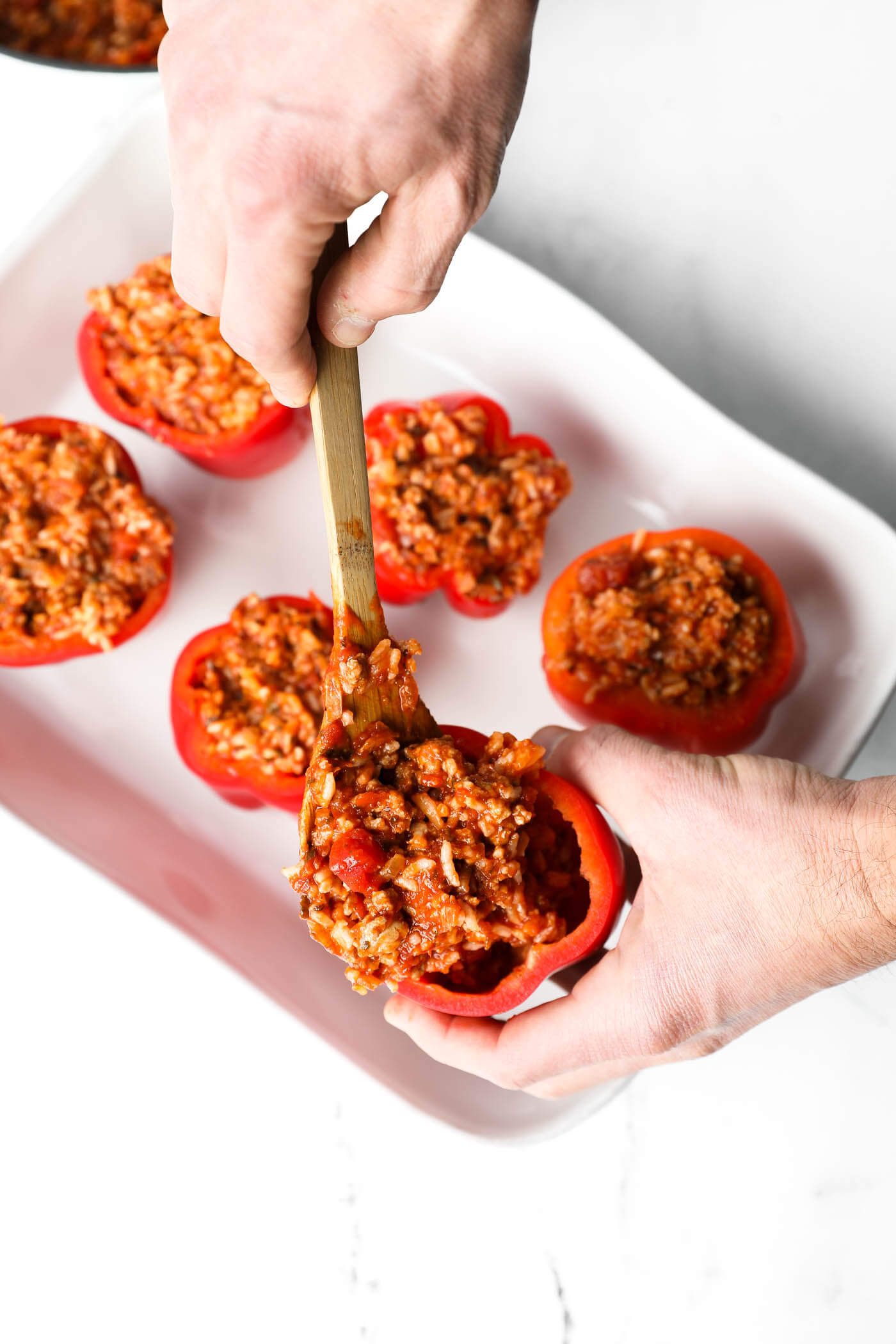 A hand holding one bell pepper and using a wooden spoon to add a scoop of the filling mixture to the uncooked pepper.