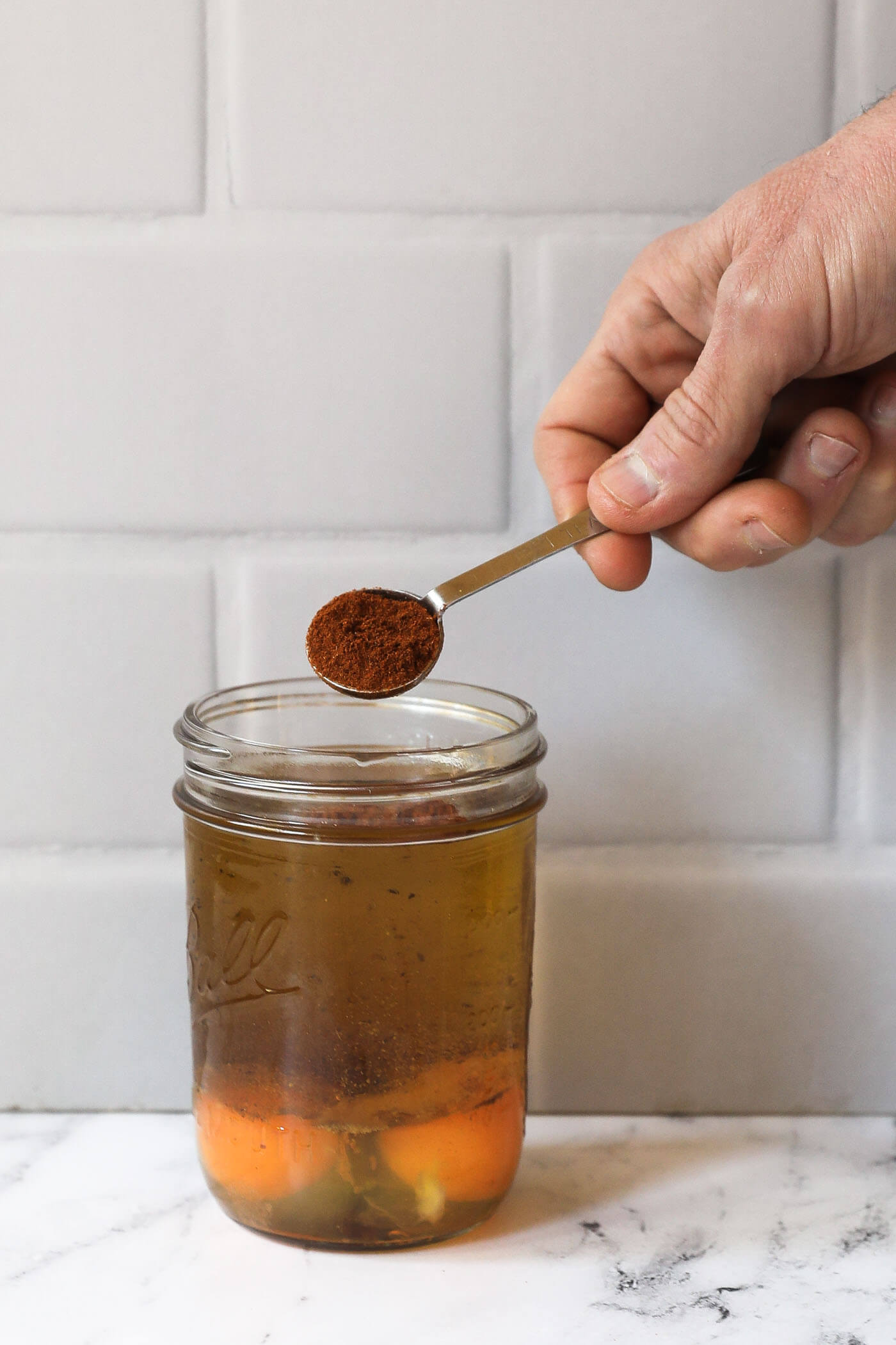 A hand pouring a teaspoon of chili seasoning into a mason jar filled with sauce ingredients.