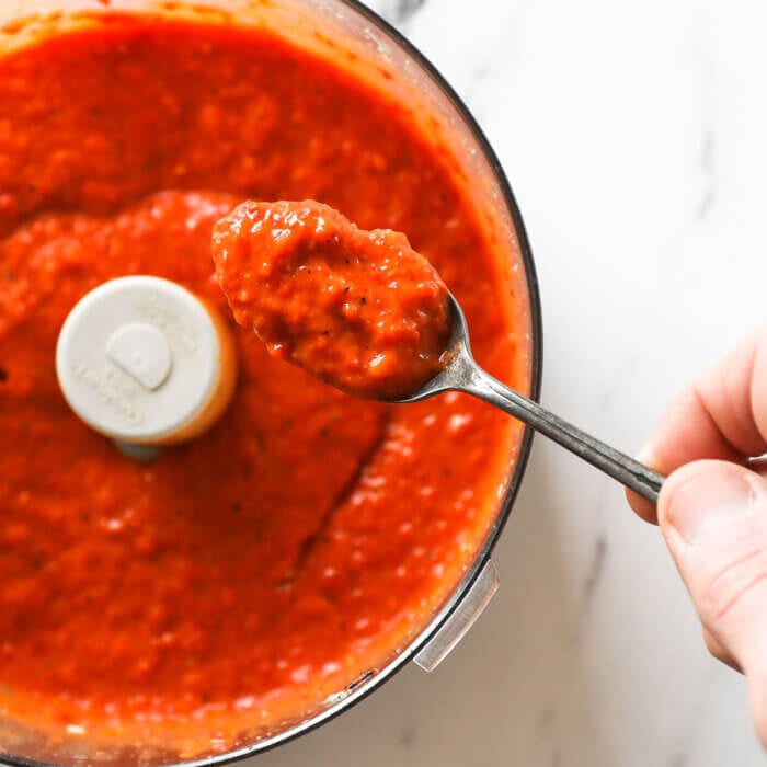 Overhead image of a spoonful of red pepper sauce over a food processor.