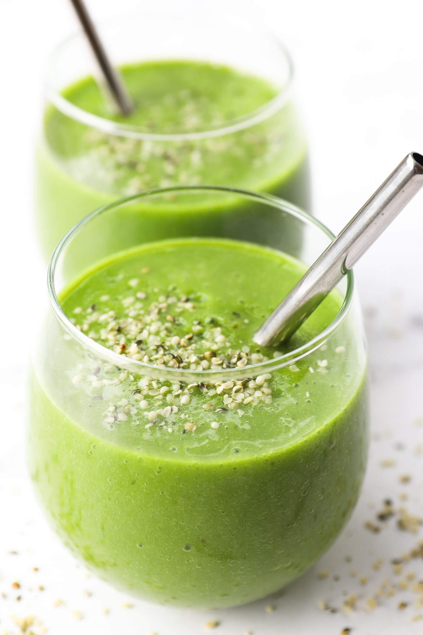 Two glasses of a spinach smoothie with straws in the glasses and hempseeds sprinkled on top.