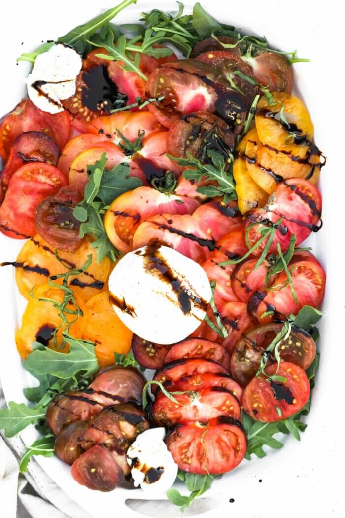 Overhead image of a platter layered with sliced heirloom. tomatoes, burrata cheese, basil, arugula and balsamic vinegar drizzled on top.