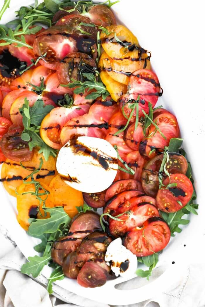 Overhead image of a platter angled to the side a bit, layered with sliced heirloom. tomatoes, burrata cheese, basil, arugula and balsamic vinegar drizzled on top.
