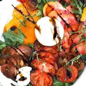 Close up overhead image of sliced heirloom tomatoes on a bed or arugula lettuce. Topped with burrata cheese, basil and drizzled with balsamic vinegar.