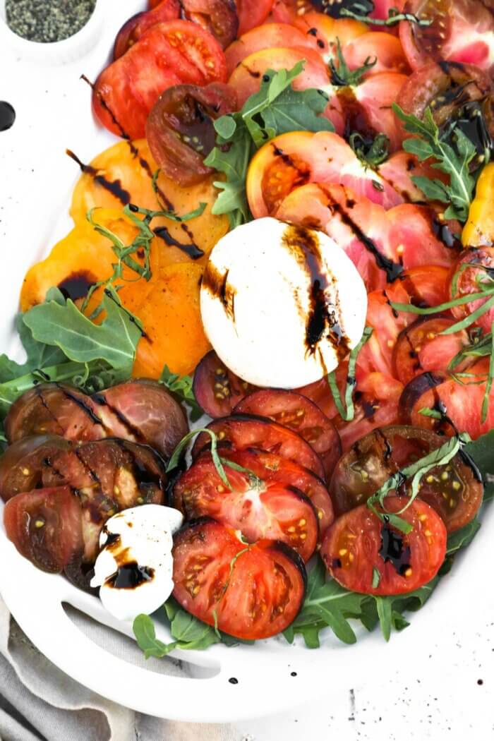 Close up overhead image of sliced heirloom tomatoes on a bed or arugula lettuce. Topped with burrata cheese, basil and drizzled with balsamic vinegar.