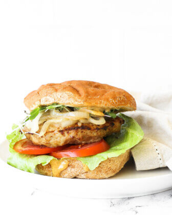 Image of a loaded turkey burger on a gluten free bun. Toppings include chipotle aioli, lettuce, tomato, turkey patty, caramelized onions and microgreens.