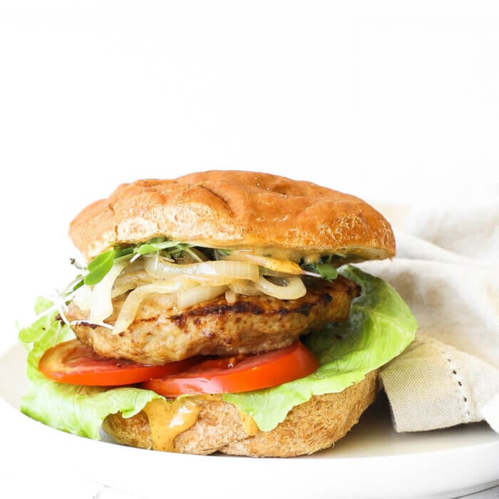 Image of a loaded turkey burger on a gluten free bun. Toppings include chipotle aioli, lettuce, tomato, turkey patty, caramelized onions and microgreens.