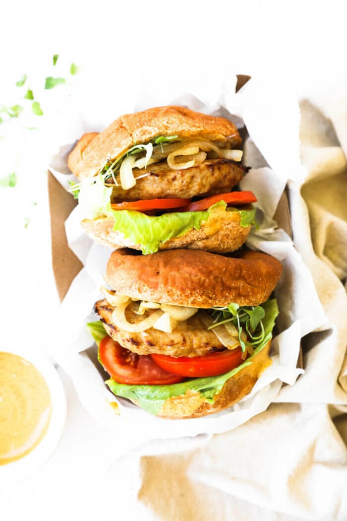 Two loaded turkey burgers in a to-go container with parchment paper. Extra chipotle aioli sauce on the side.