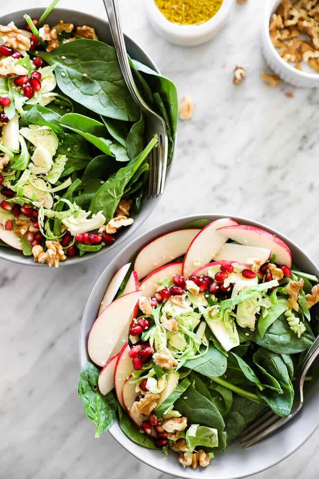 Vertical image of apple walnut spinach salad in two bowls with forks dug in. Dressing and walnuts on the side. 