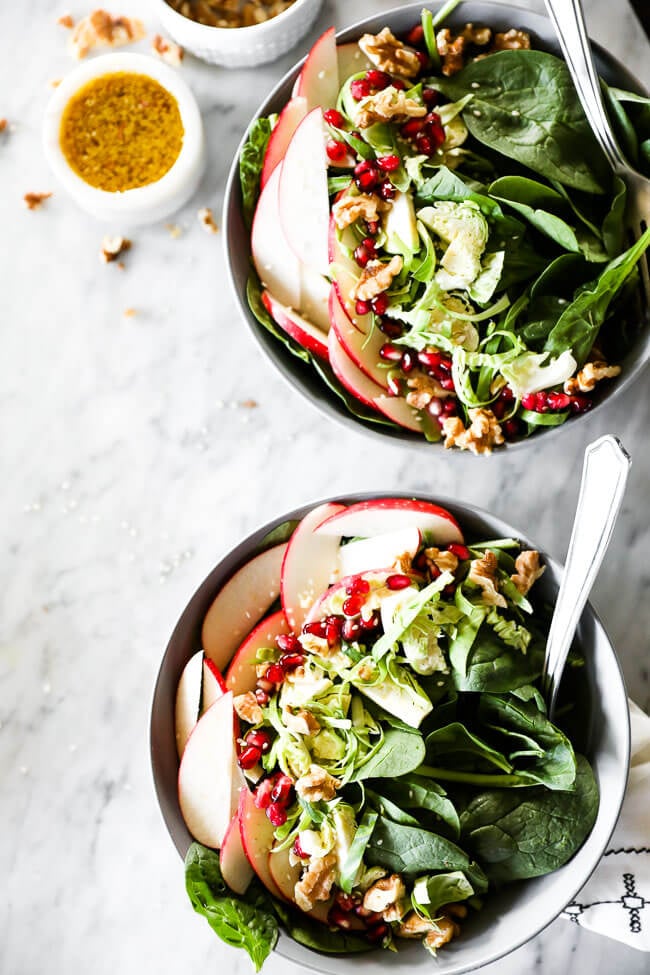 Vertical image of apple walnut spinach salad in two bowls with forks dug in. Dressing on the side. 