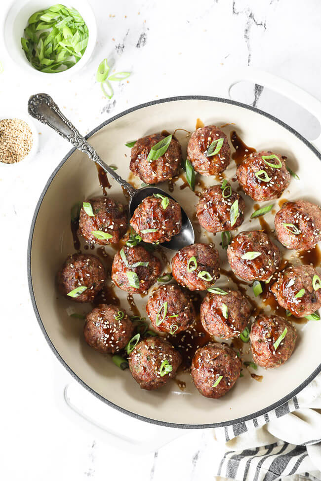 Overhead vertical image of asian meatballs in a skillet with serving spoon. Drizzled with sauce and topped with chopped green onion and sesame seeds. 