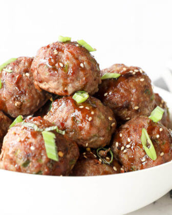 Vertical angled close up image of asian meatballs in a bowl with sauce, green onion and sesame seeds on top.