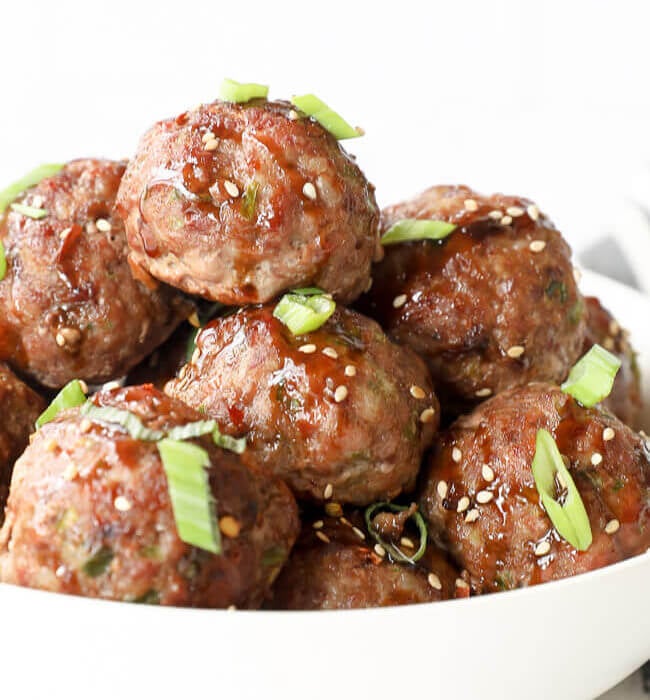 Vertical angled close up image of asian meatballs in a bowl with sauce, green onion and sesame seeds on top.