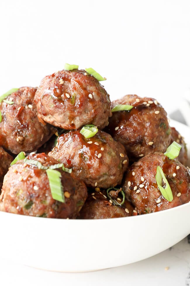 Vertical angled close up image of asian meatballs in a bowl with sauce, green onion and sesame seeds on top. 