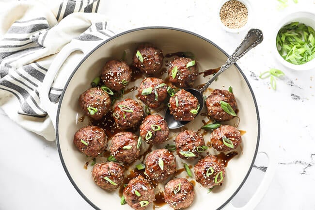 Immagine orizzontale di polpette asiatiche in una padella con cucchiaio di servizio. Condite con salsa e condite con cipolla verde tritata e semi di sesamo. 