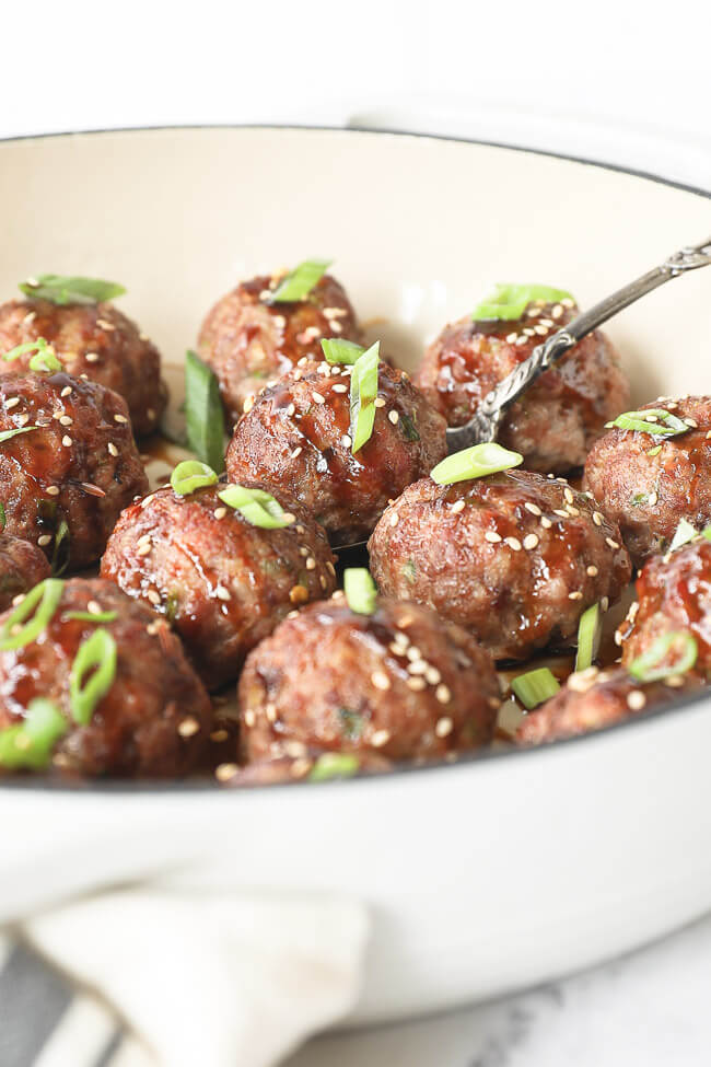 Vertical angled image of asian meatballs in a skillet with serving spoon. Drizzled with sauce and topped with chopped green onion and sesame seeds. 