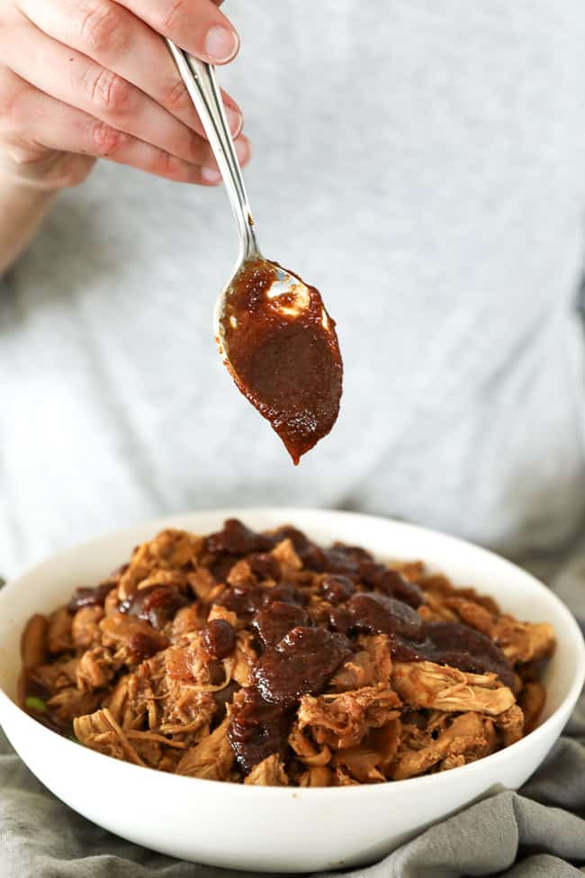 Vertical straight on image of drizzling BBQ sauce on top of bowl of BBQ pulled chicken. 