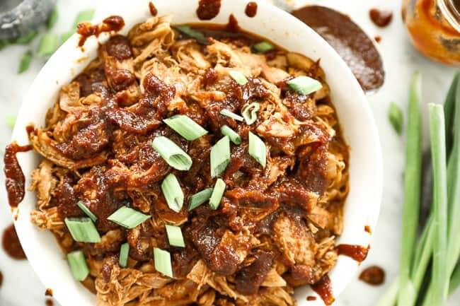 Horizontal overhead image of BBQ pulled chicken in bowl with extra sauced drizzled everywhere. Topped with chopped green onion. 