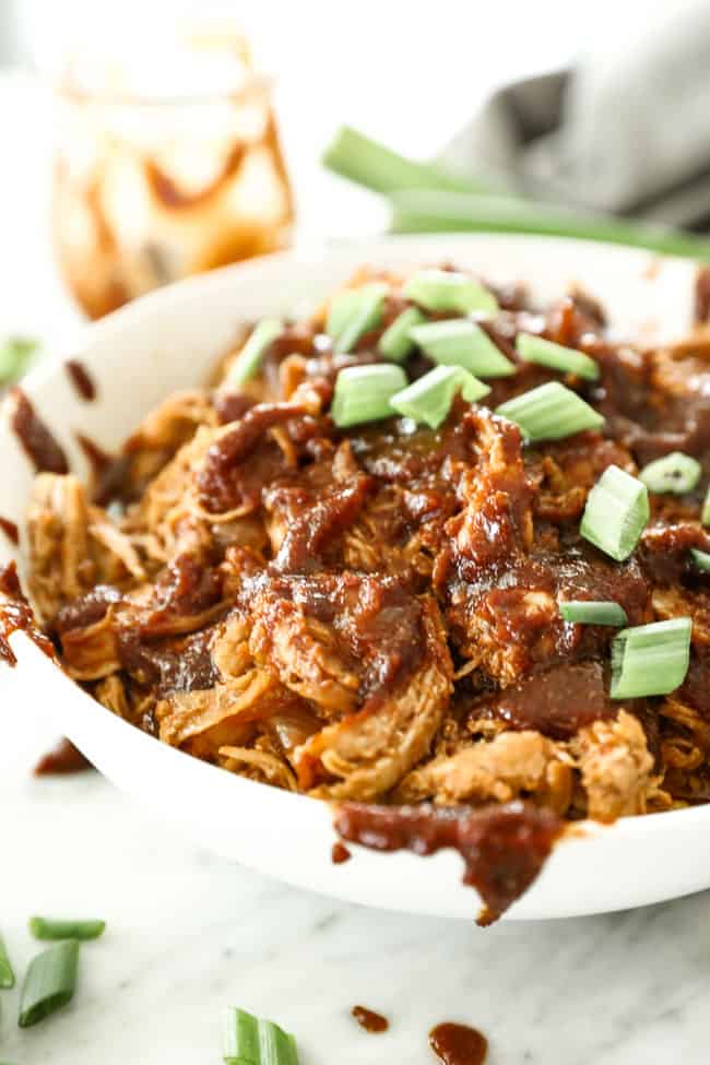 Angled vertical image of BBQ pulled chicken in a bowl with lots of extra sauce drizzled everywhere and topped with chopped green onion. 