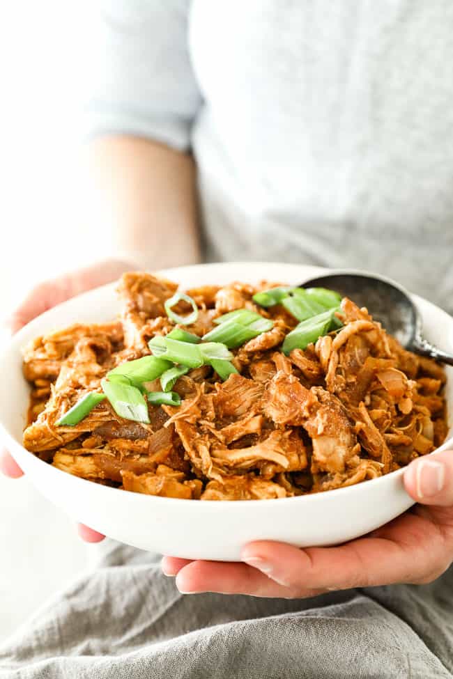 Angled vertical image of holding bowl of BBQ pulled chicken with chopped green onion on top. 