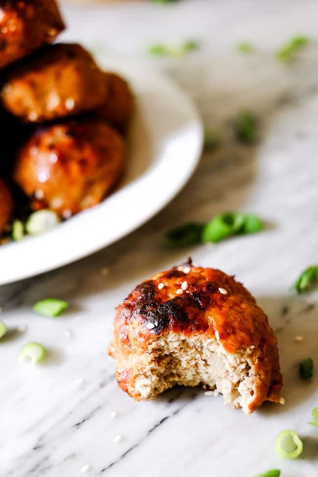 Baked turkey meatballs on a plate in the background with one meatball with a bite take out of it in the foreground. Chopped green onions and sesame seeds in the background. 