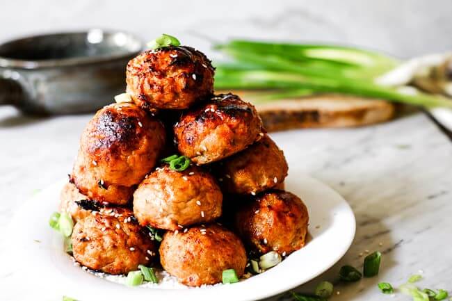 Baked turkey meatballs stacked on a plate with chopped green onion and sesame seeds on top. Green onion and a serving bowl in the background. 