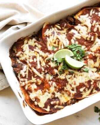 A baking dish filled with cheesy beef enchiladas topped with melted cheese, chopped cilantro, and lime wedges on a white surface.