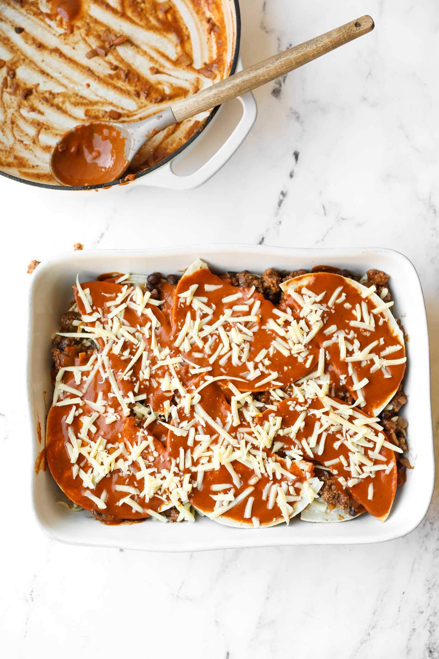 A white baking dish filled with a layered casserole topped with sauce and shredded cheese sits on a marble surface; an empty pot with sauce residue is beside it.