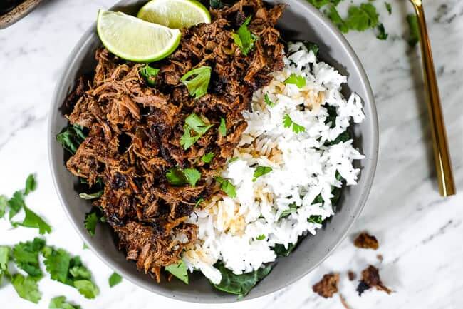 Beef barbacoa in a bowl with greens and rice and topped with chopped cilantro and lime wedges. 