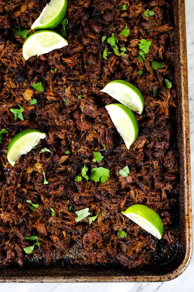 Beef barbacoa on a sheet pan topped with chopped cilantro and lime wedges. 