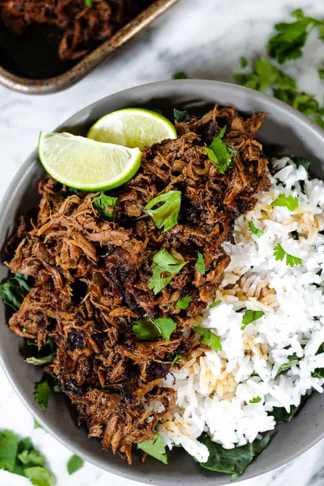 Beef barbacoa in a bowl with greens and rice and topped with chopped cilantro and lime wedges. 