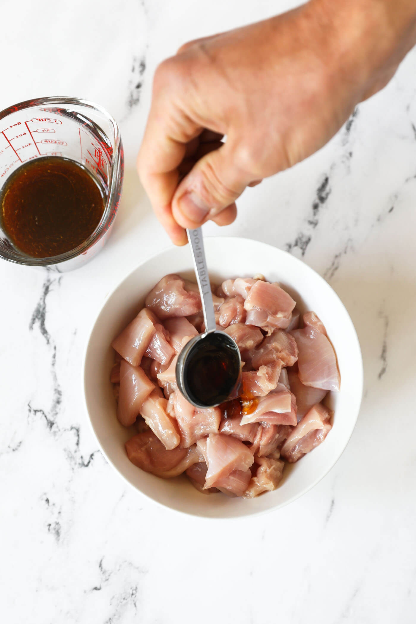 A hand spooning sauce over a bowl of raw chicken thighs cut up into bite sized pieces.