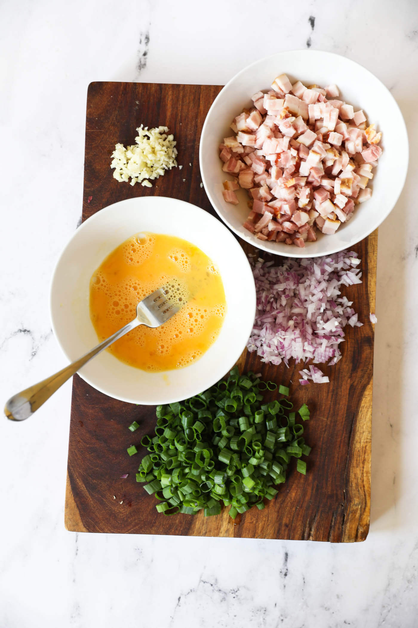 Overhead image of prepped ingredients on a cutting board - minced garlic, chopped bacon in a bowl, chopped shallot, chopped green onion and whisked egg in a bowl.