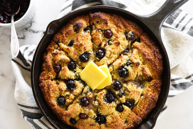 Horizontal overhead image of blueberry cornbread in a cast iron skillet with grass fed butter melting on top. 