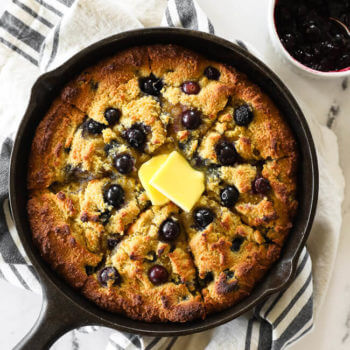 Blueberry cornbread in a skillet with blueberry sauce on the side and grass fed butter melting on top.