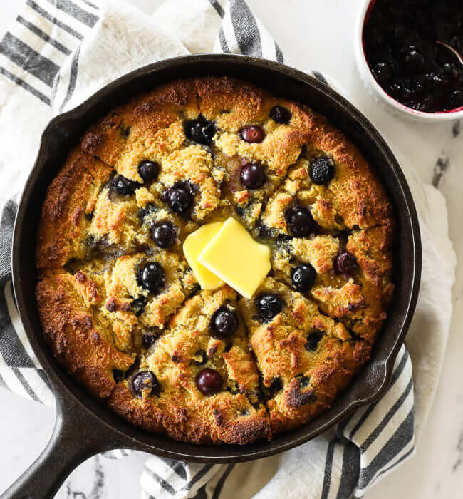 Blueberry cornbread in a skillet with blueberry sauce on the side and grass fed butter melting on top.
