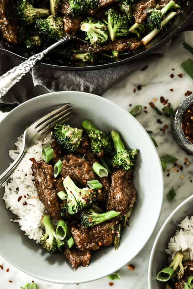 Broccoli beef stir fry served in a bowl with white rice and topped with chopped green onion and crushed red pepper. 