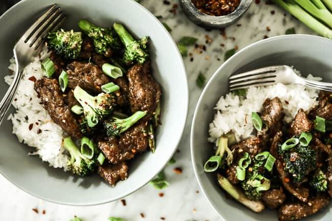 Broccoli beef stir fry served up in two bowls with white rice, crushed red pepper sprinkled on top and forks in the bowls. 