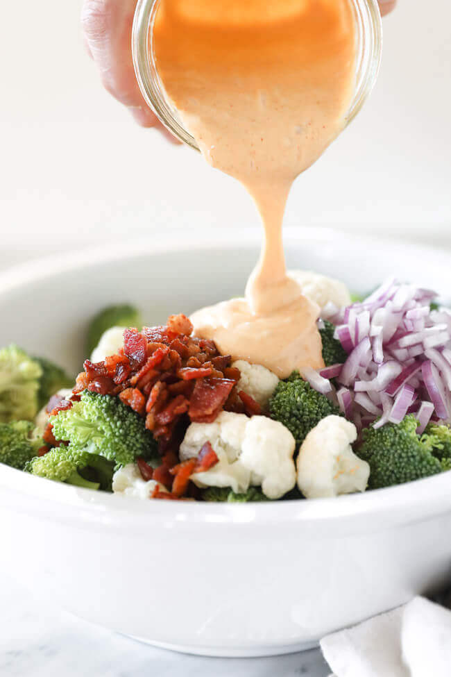 Broccoli and cauliflower florets in a bowl with a pile of bacon and red onion. Sauce being poured from top.